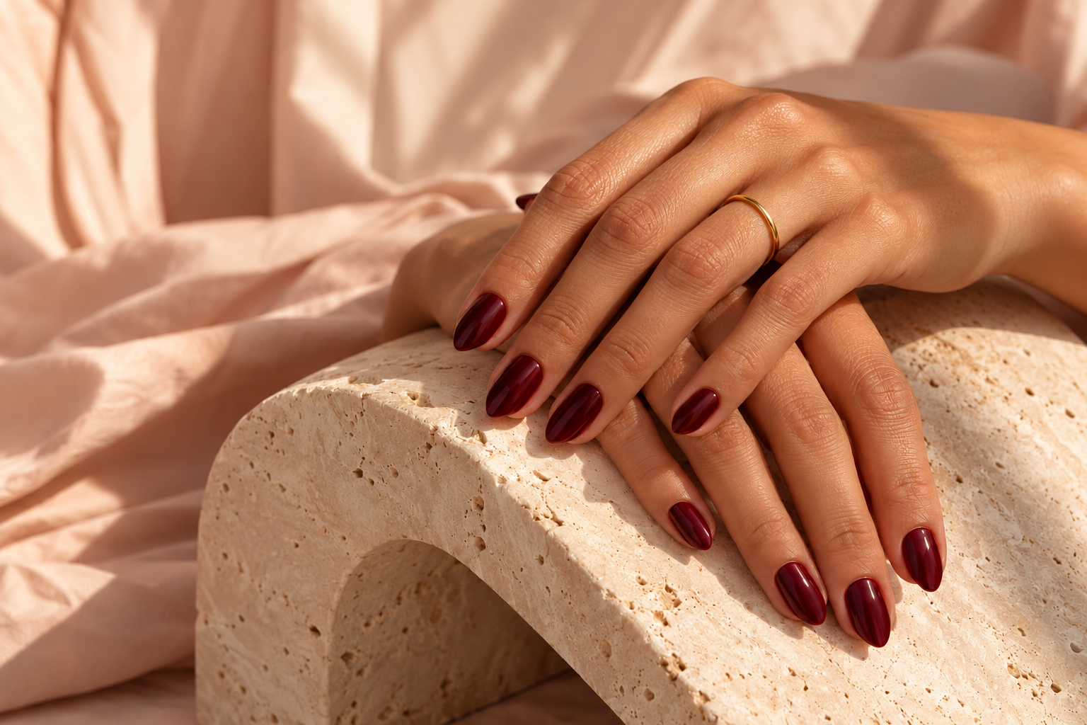 Hands with glossy burgundy nails resting on warm travertine